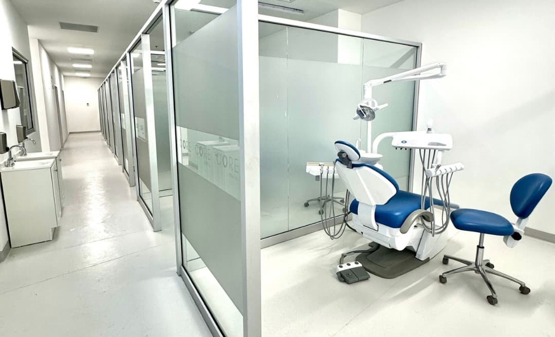 Dental treatment room with a blue dental chair, overhead lamp and instrument tray behind a glass partition