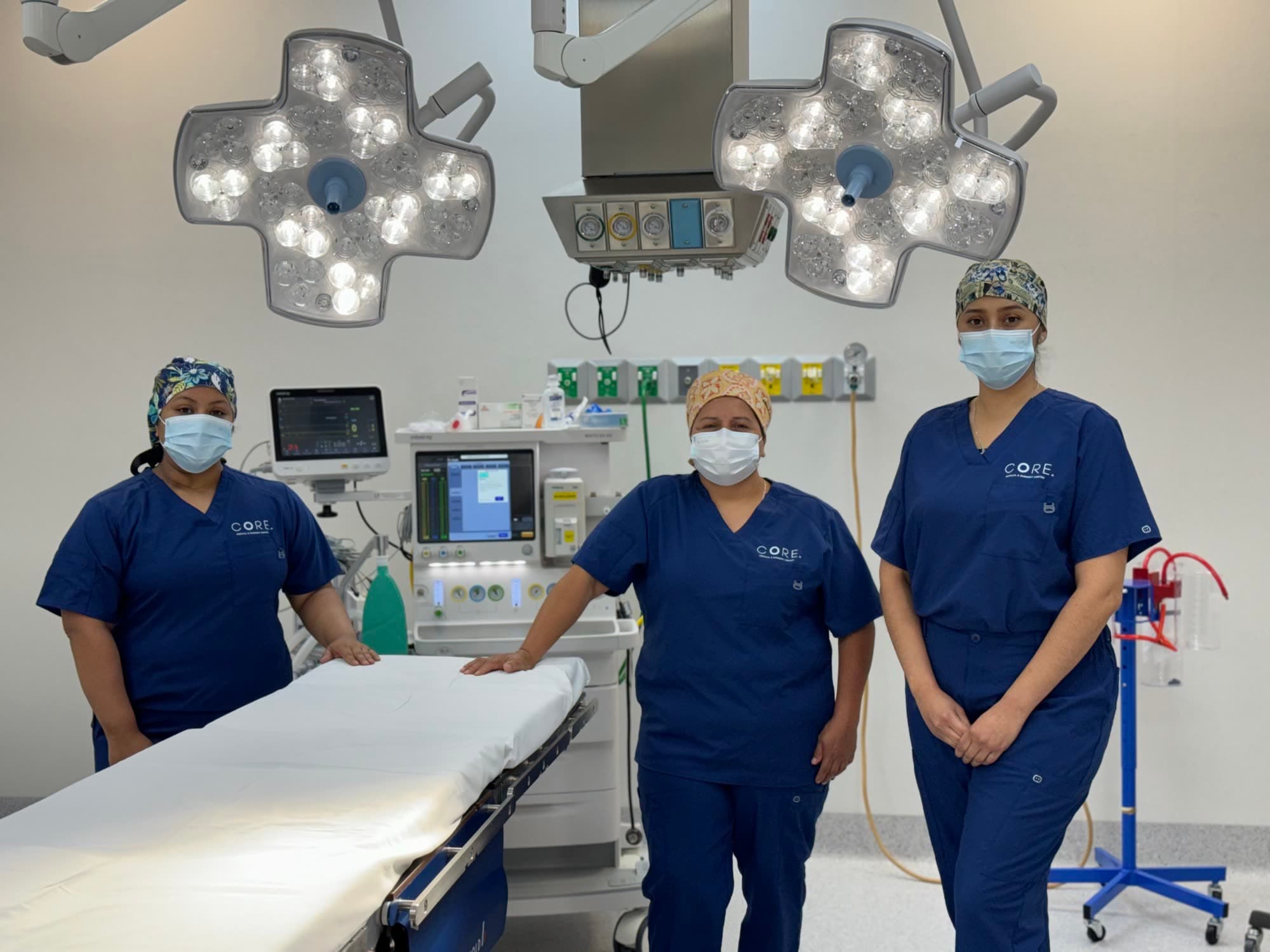 Tres integrantes del equipo quirúrgico de CORE con pijama quirúrgica azul marino de CORE, gorro y cubrebocas, de pie junto a una mesa de operaciones bajo lámparas quirúrgicas