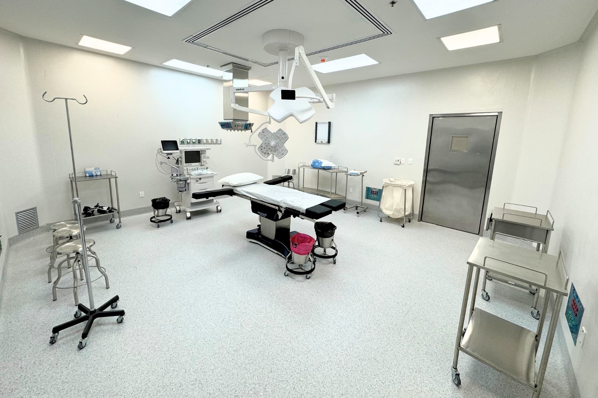 Empty operating room with a surgical table, overhead surgical lights, an anesthesia machine and stainless steel doors
