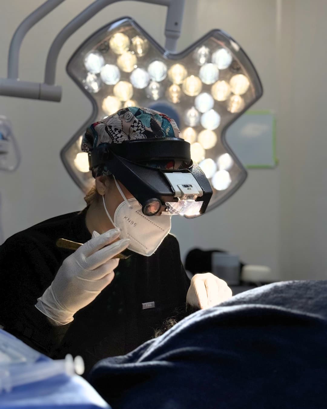 A surgeon wearing magnifying loupes and a headlamp works beneath a surgical light over a draped patient
