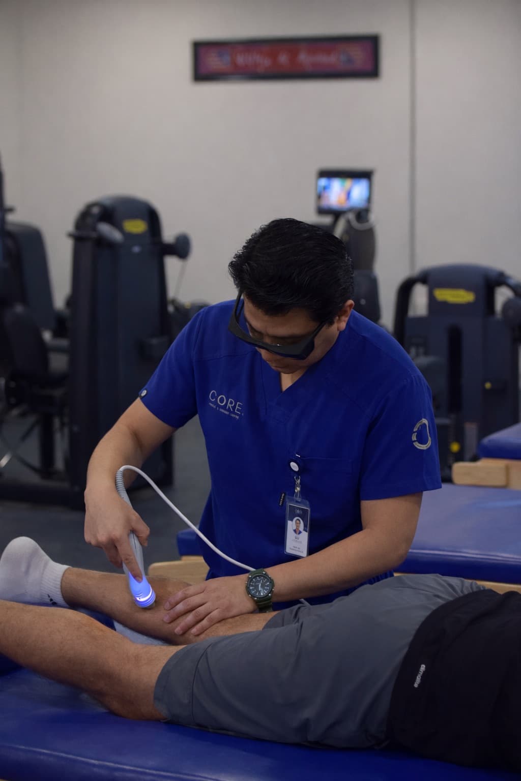 A therapist in blue scrubs applies a handheld laser device to a patient's lower leg on a treatment table
